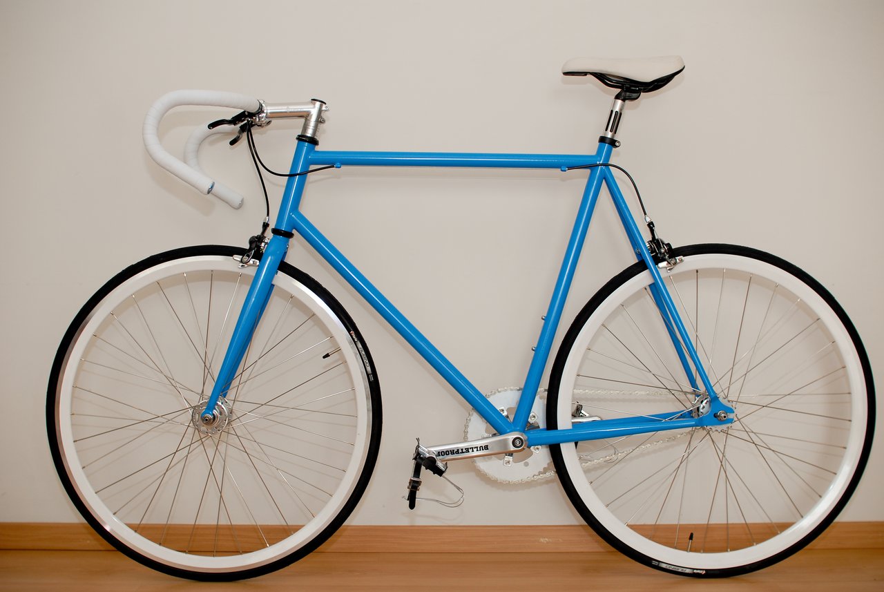 A blue road bike with white handlebars and wheels leans against a plain wall indoors.