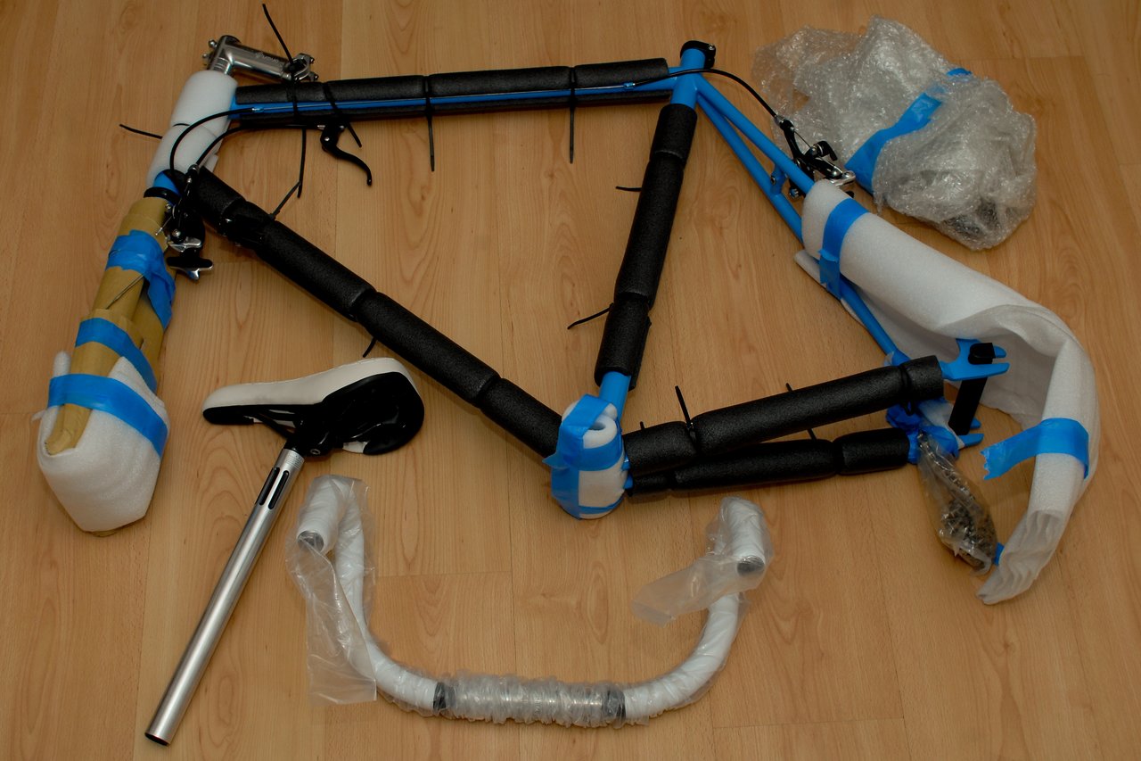 Partially assembled blue bicycle frame with protective padding, handlebars, and seat post laid out on a wooden floor.