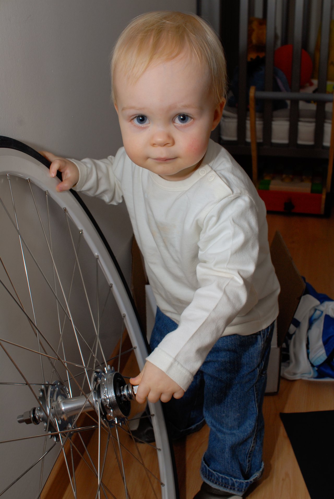 A young child in a white shirt and jeans holds a large bicycle wheel while standing indoors.