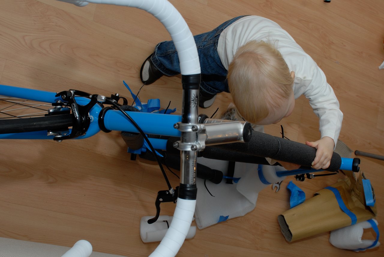 A child in a white shirt removes protective wrapping from a partially assembled blue bicycle on a wooden floor.