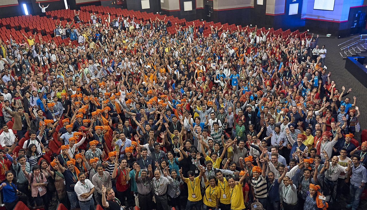 A large group of people in an auditorium cheer and raise their hands, some wearing orange headbands.