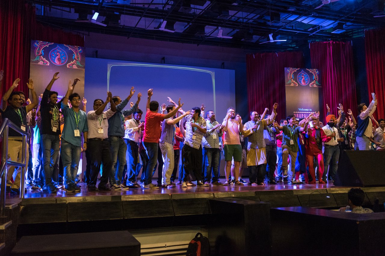 A large group of people on stage raising their hands and singing or cheering together at an event.