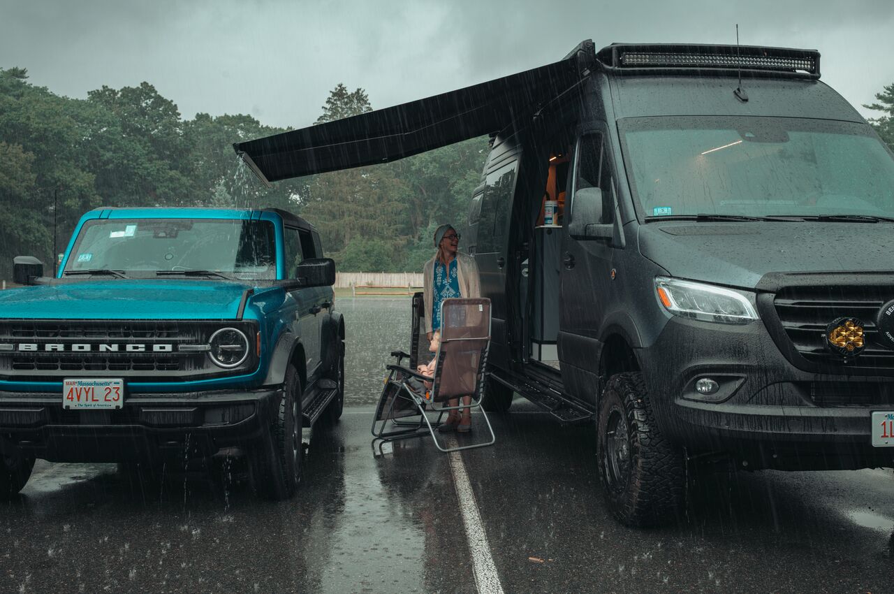 A woman and child stay dry under the awning of a black camper van, parked next to a blue Ford Bronco on a rainy day.