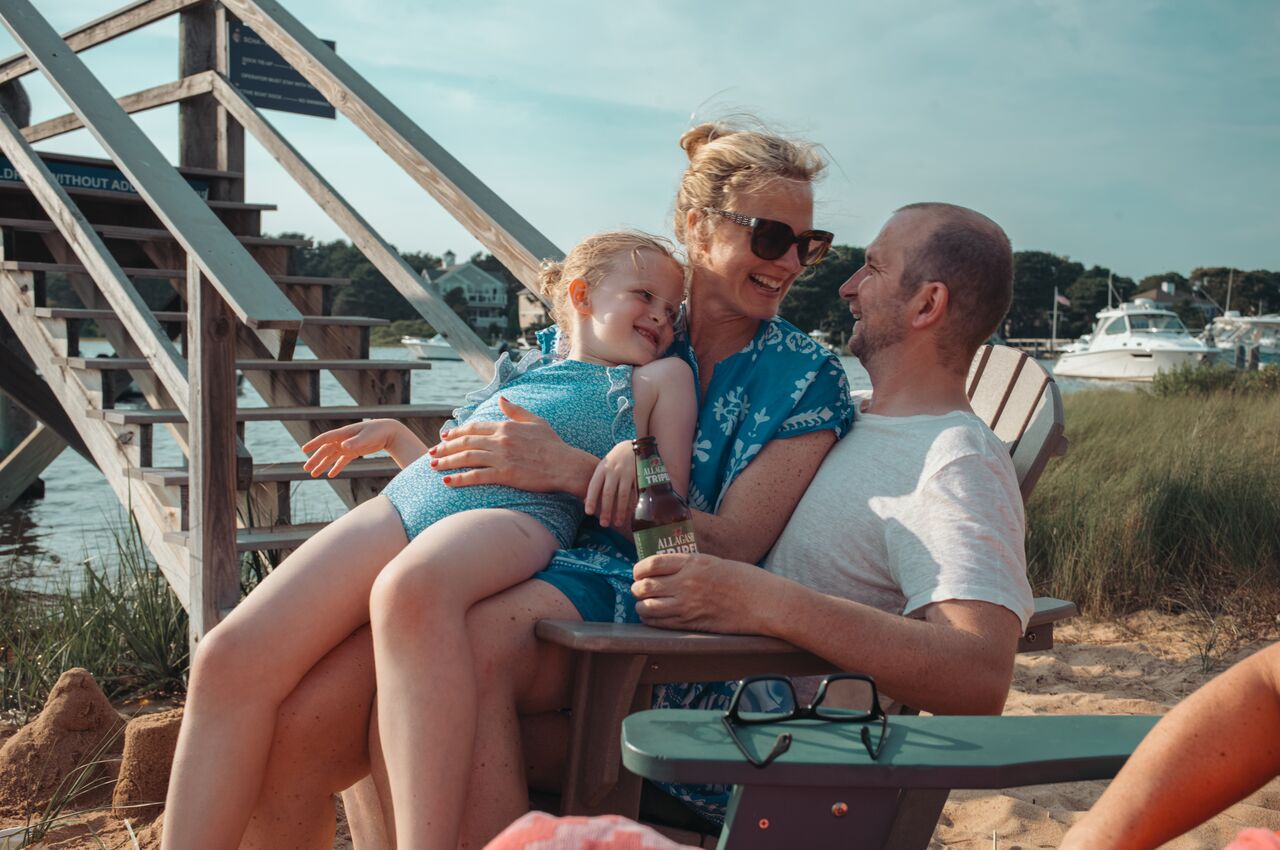 A woman sits on a man's lap holding a young girl, all smiling and relaxed on the beach near wooden stairs and water.