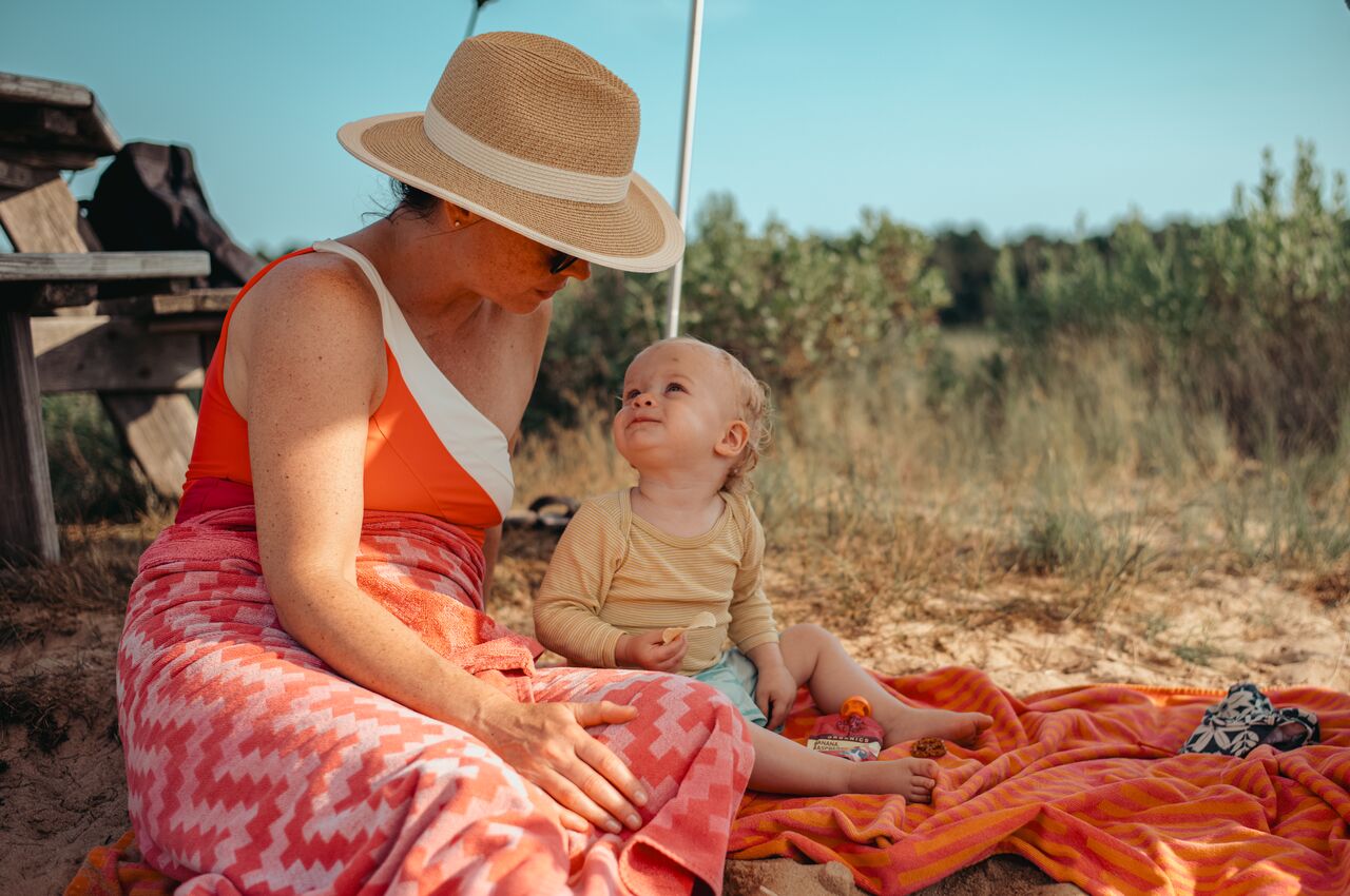 A woman in a sunhat sits on a beach blanket with a smiling toddler, surrounded by sand and grassy dunes.