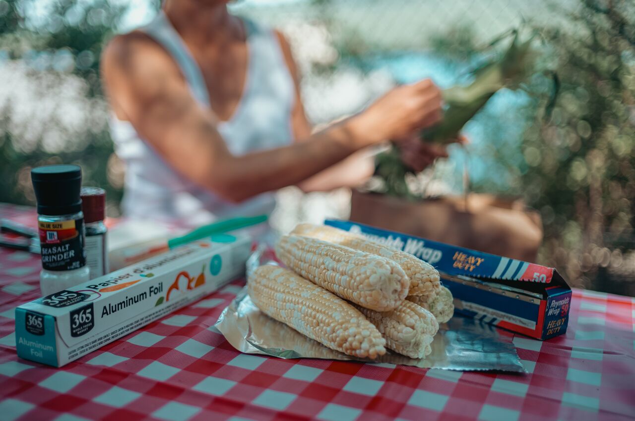 Close-up of uncooked corn on the cob resting on aluminum foil, with a person in the background husking more corn at a picnic table.