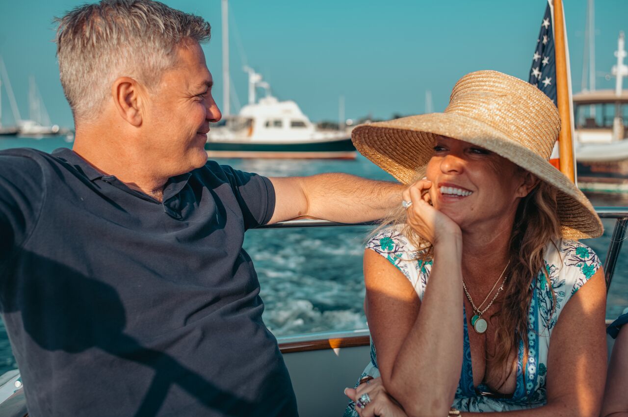 A man and woman sit closely together on a boat, smiling and looking at each other while enjoying a sunny day on the water.