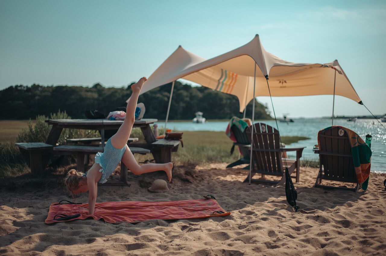 A young girl does a cartwheel on the beach, with a shaded picnic area and a calm bay with boats in the background.