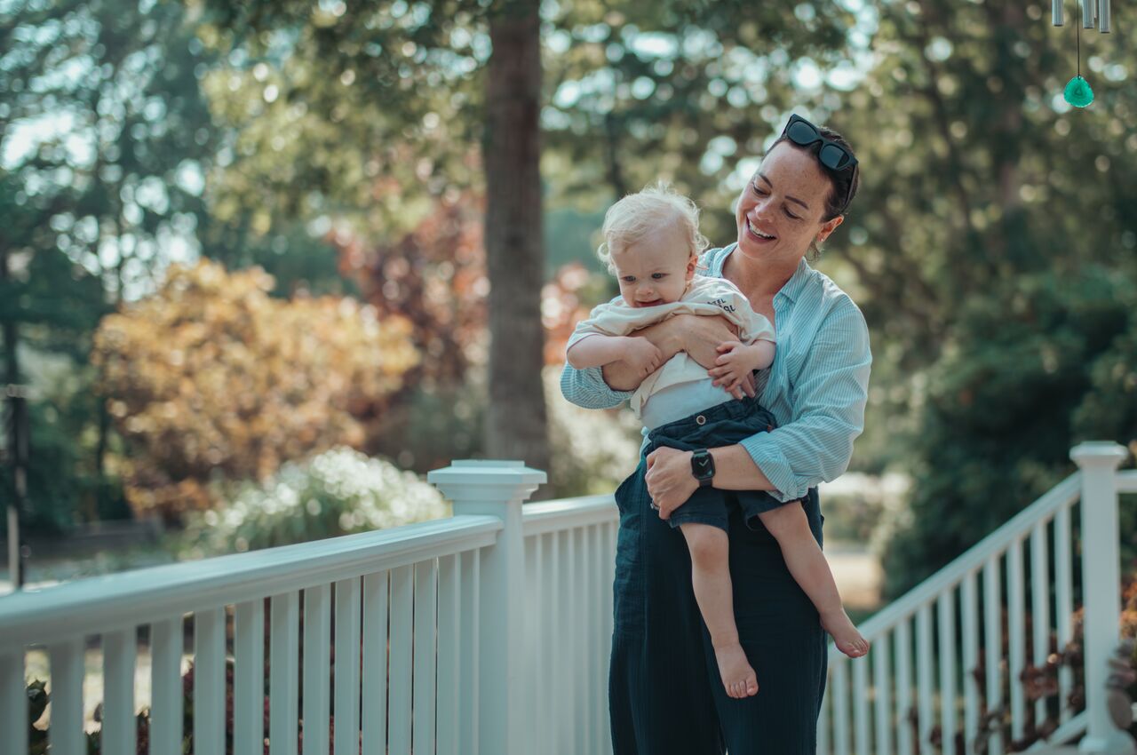 A woman holds a smiling toddler in her arms while standing on a porch, both appearing happy and relaxed outdoors.
