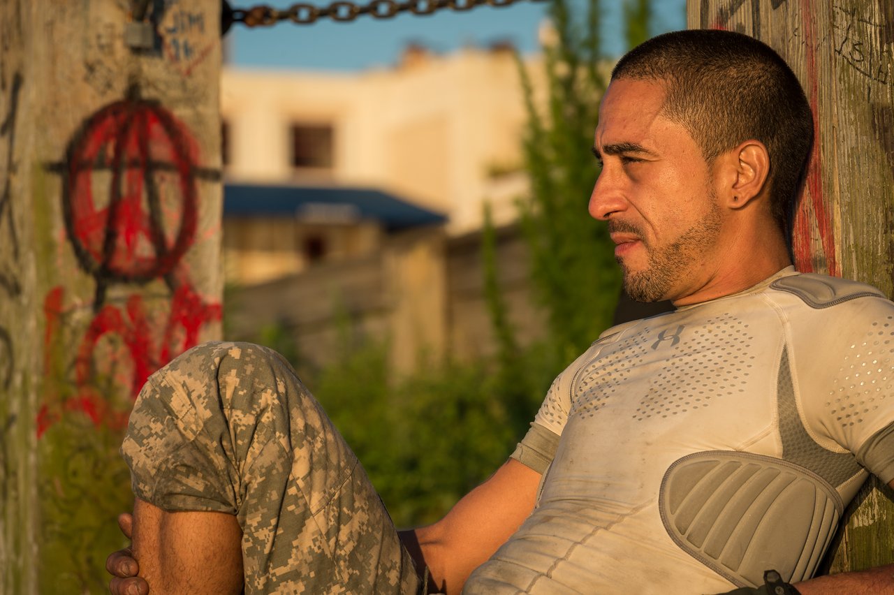 A man in tactical clothing sits against a graffiti-covered post, holding a gun in his hand.