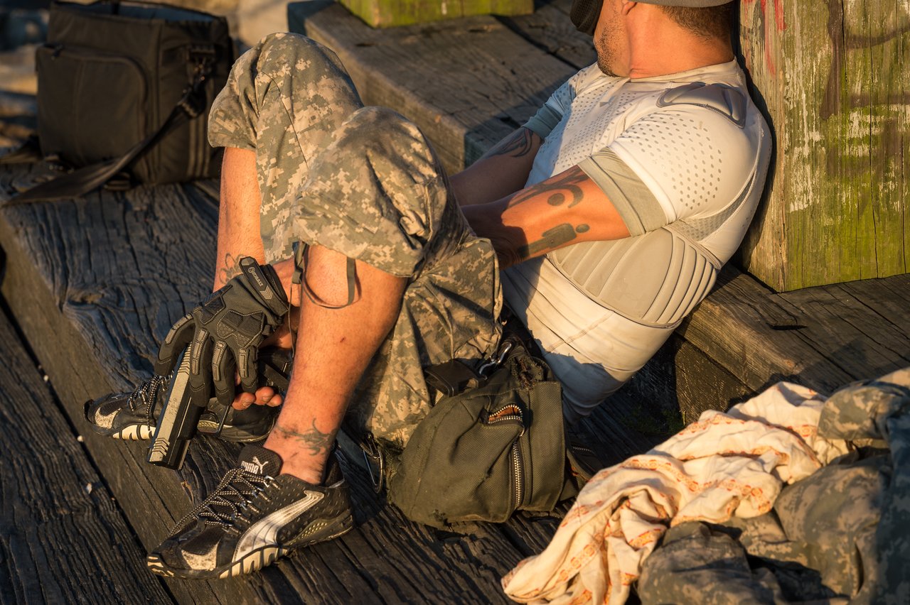 A person in camouflage pants sits on a wooden surface, holding a handgun with a gloved hand.