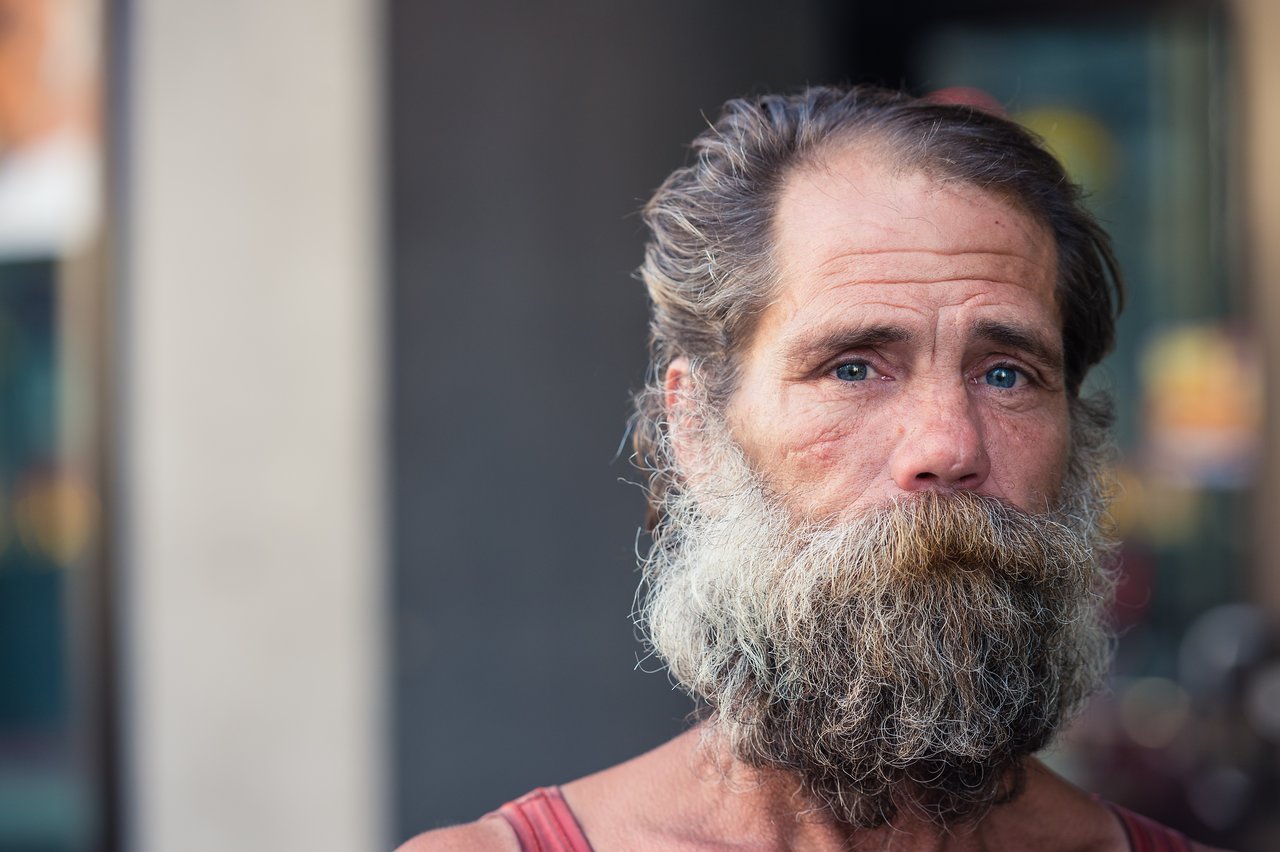 A bearded man with blue eyes looks directly at the camera on a city street.