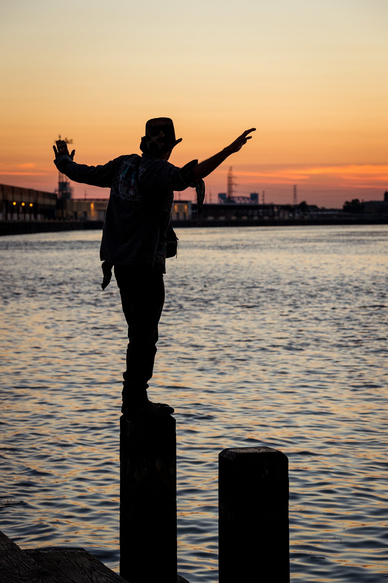 A person balances on a wooden post by the water at sunrise, arms outstretched.