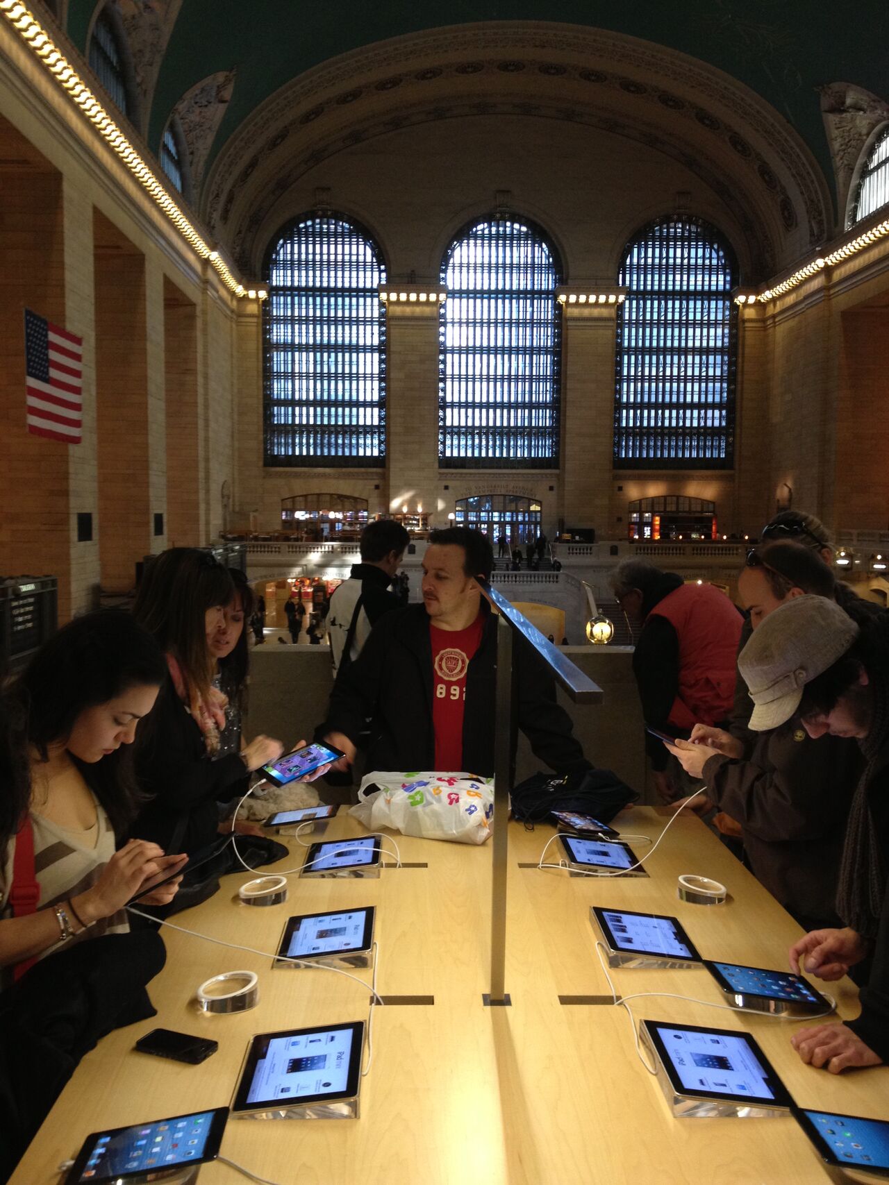 People trying iPads at a long table in the Apple Store on a balcony of Grand Central Terminal, with the concourse's arched windows behind them.