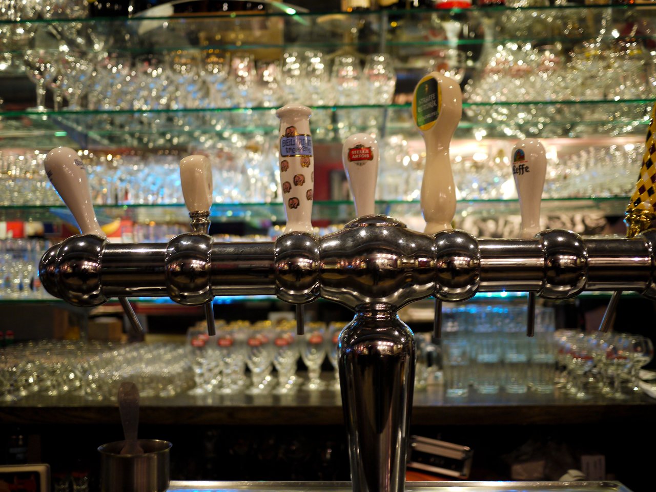 Chrome beer taps with Belgian beer handles, including Leffe, in front of glass shelves stacked with beer glasses.