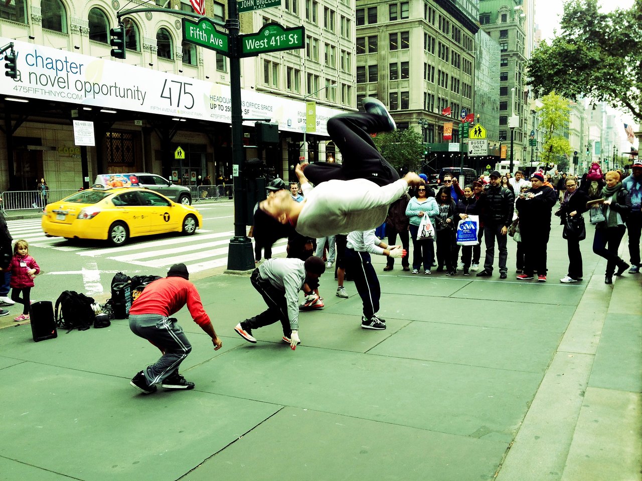 A street dancer flips in midair at Fifth Avenue and West 41st Street while a crowd watches.