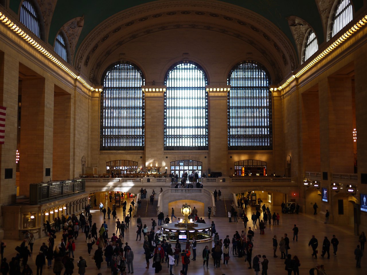 The main concourse of Grand Central Terminal, with its tall arched windows, the clock on the information booth and travelers crossing the floor.