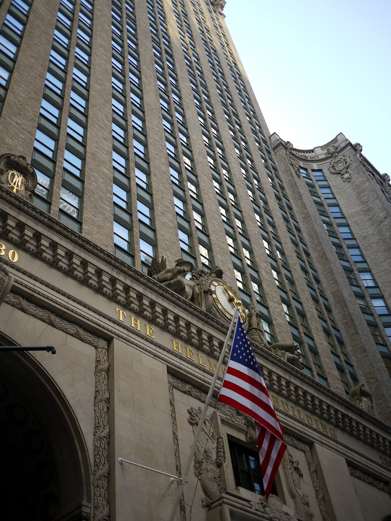 Looking up at the Helmsley Building, with an American flag over the entrance and a gilded clock and sculptures at the base of the tower.
