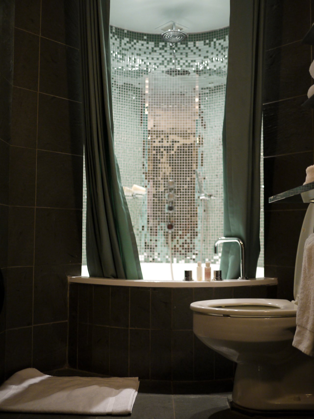 A dark-tiled hotel bathroom with a bathtub in a curtained alcove and a glass mosaic shower wall behind it.