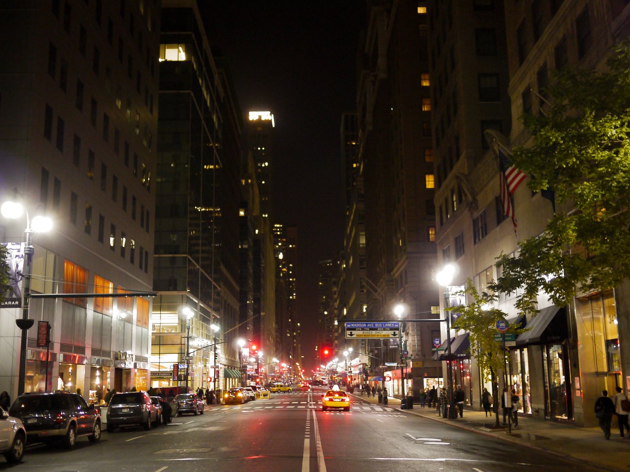 A Manhattan avenue at night, lined with lit skyscrapers and streetlights, with a yellow taxi driving toward the camera.