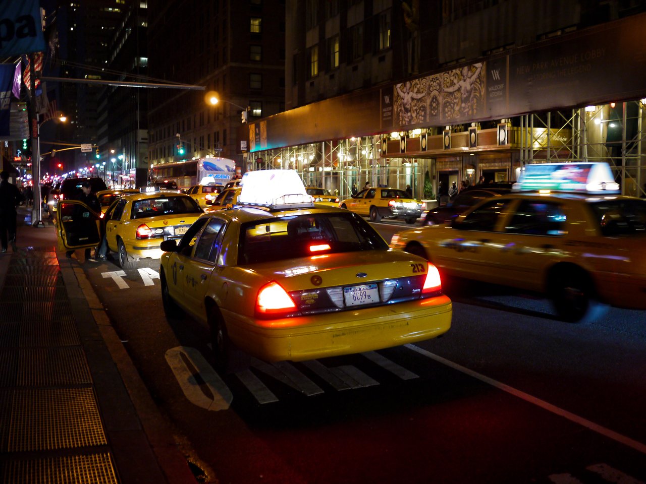 Yellow taxis at the curb on a Manhattan street at night, one with its door open as a passenger gets in.