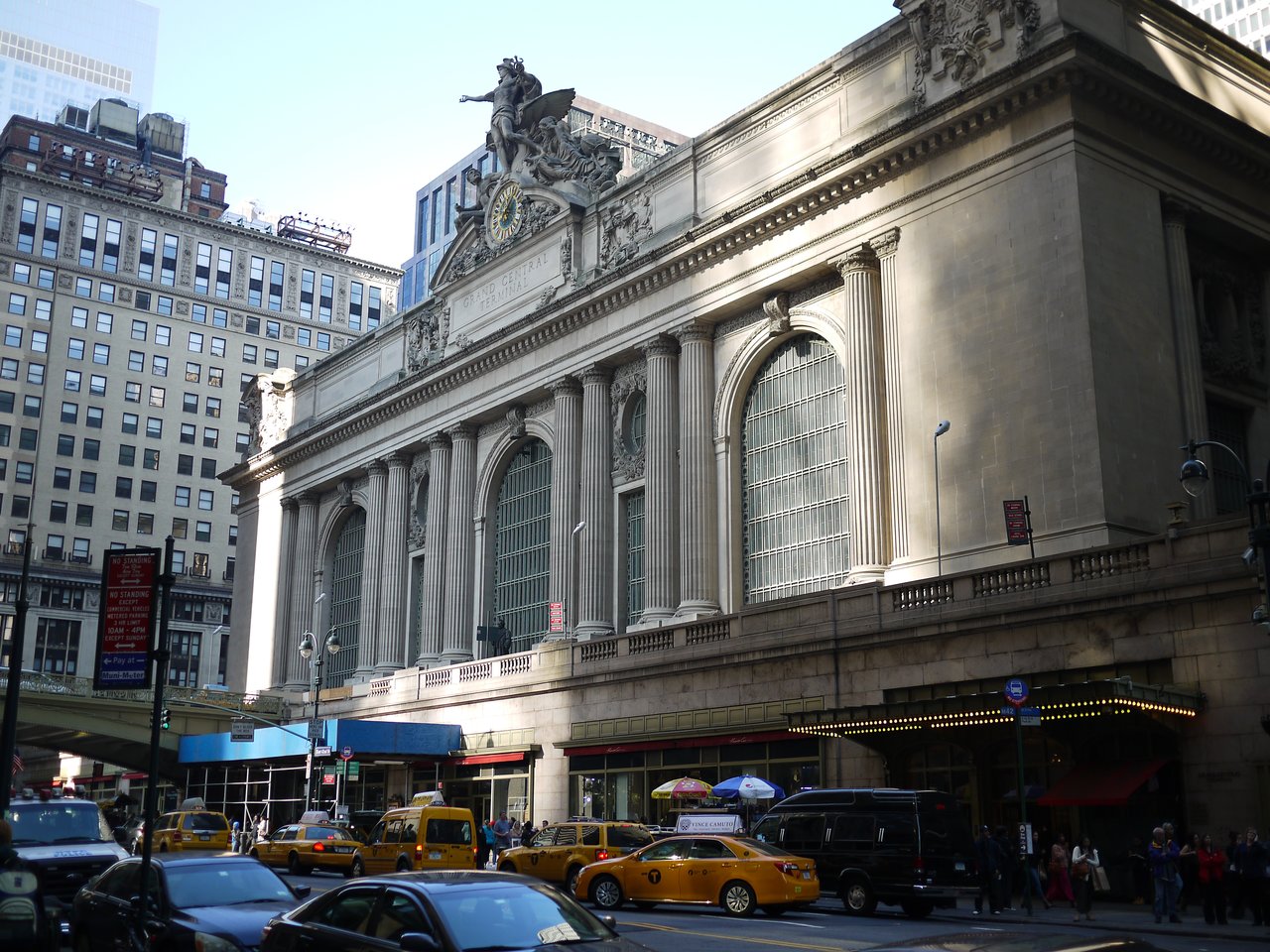 The facade of Grand Central Terminal on 42nd Street, with tall arched windows and a clock and sculptures above the entrance, and yellow taxis in front.