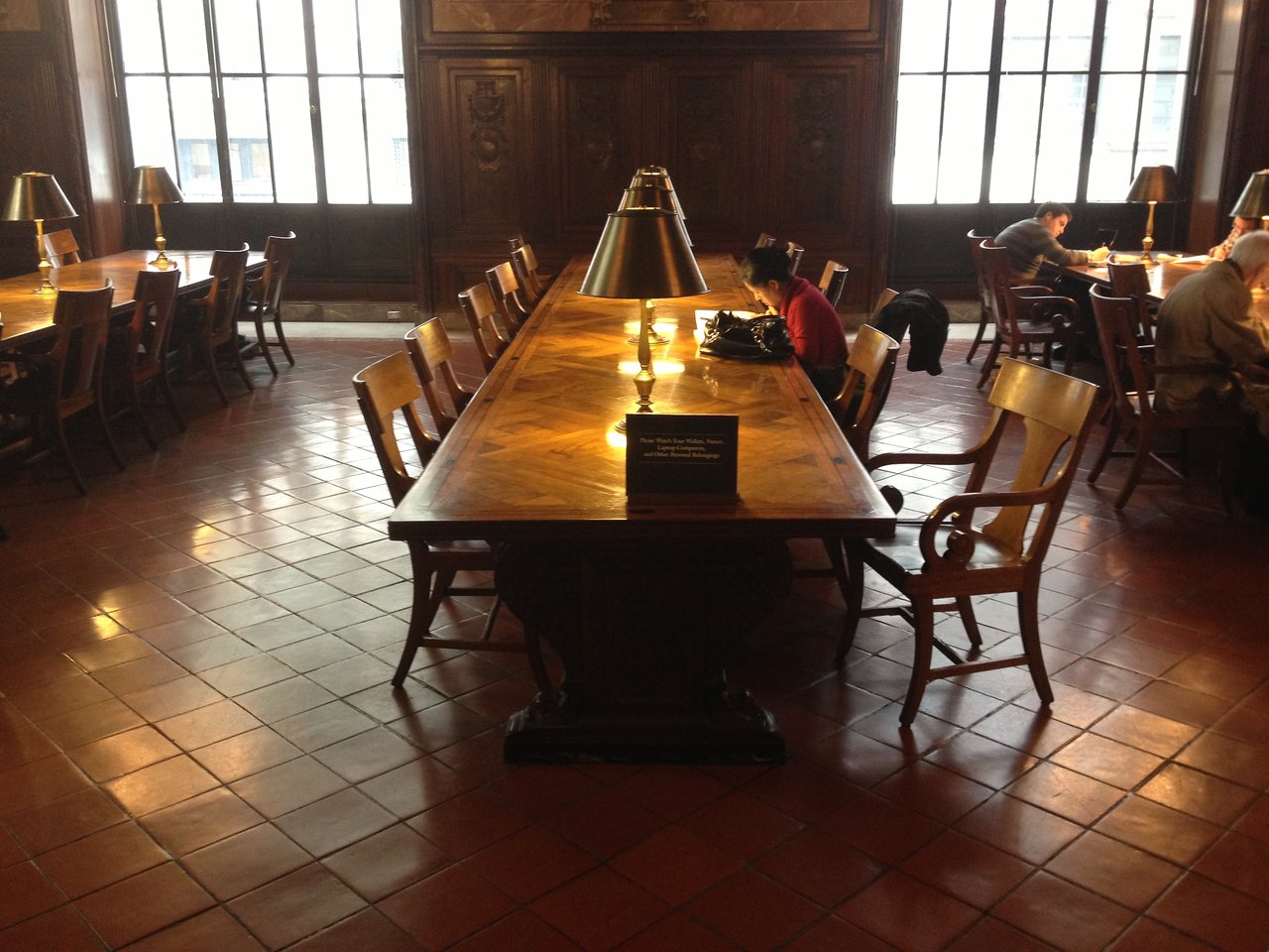 A quiet library reading room with long wooden tables, brass lamps and tall windows, with a few people reading.