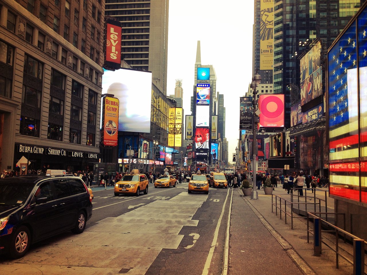 Times Square with yellow taxis, crowds and billboards, including a large American flag display on the right.