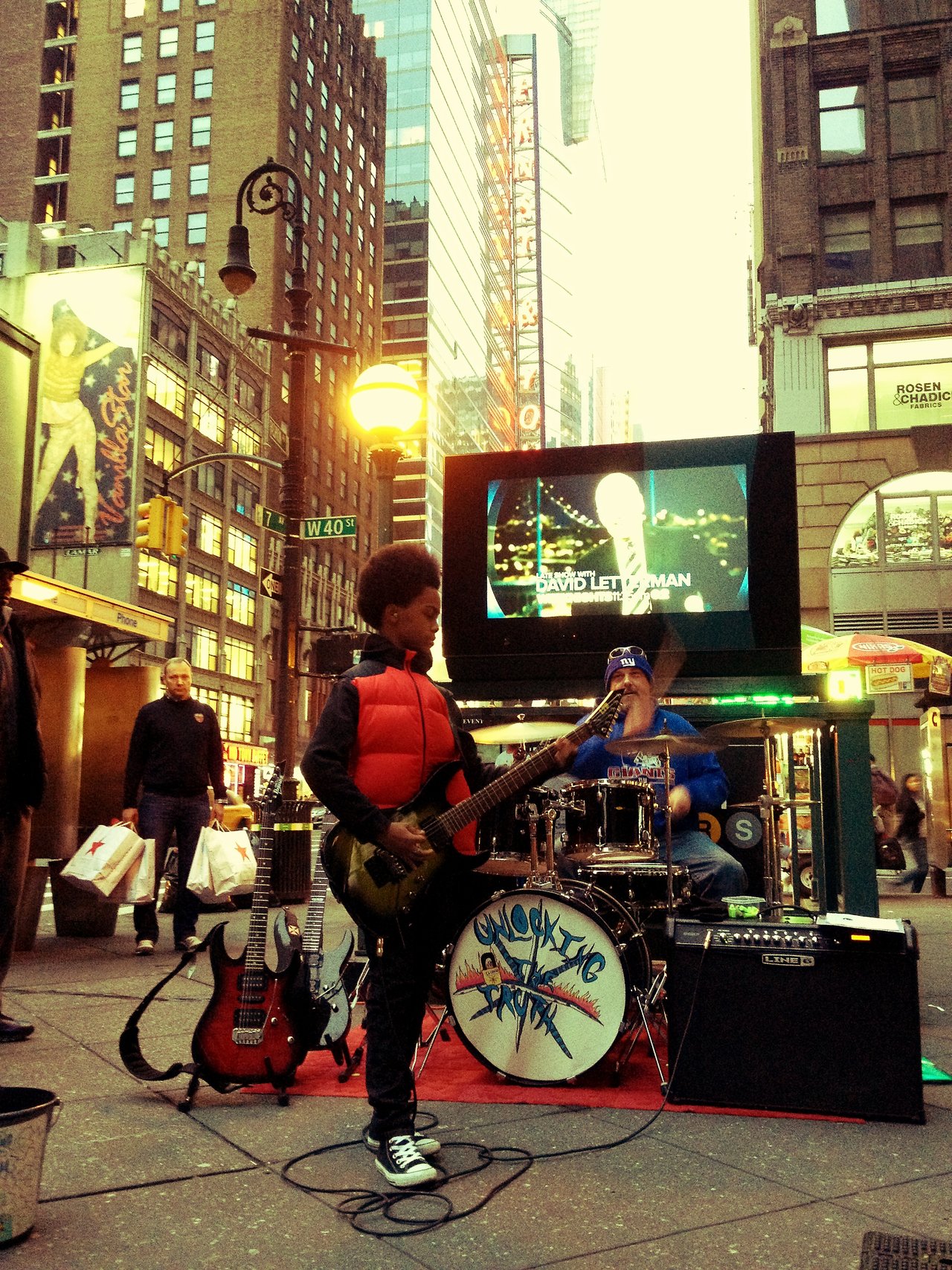 A young guitarist and a drummer playing on the sidewalk at West 40th Street, with spare guitars and an amplifier on a red rug.