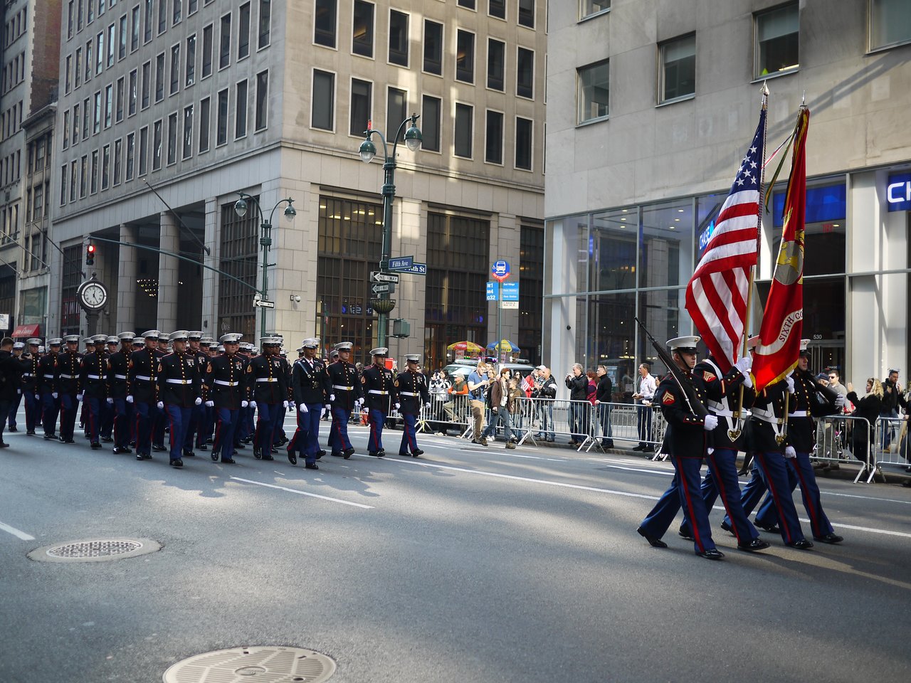 Marines in dress blue uniforms march down Fifth Avenue in the Veterans Day parade, led by the American flag and a red Marine Corps flag.