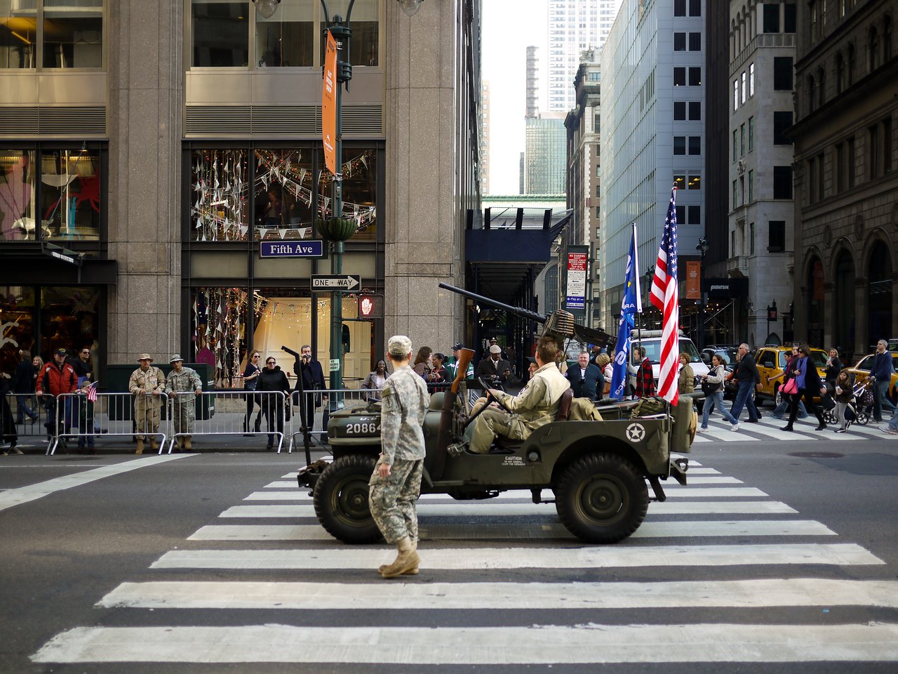 A man in camouflage walks beside a vintage army jeep carrying American flags across a Fifth Avenue crosswalk.