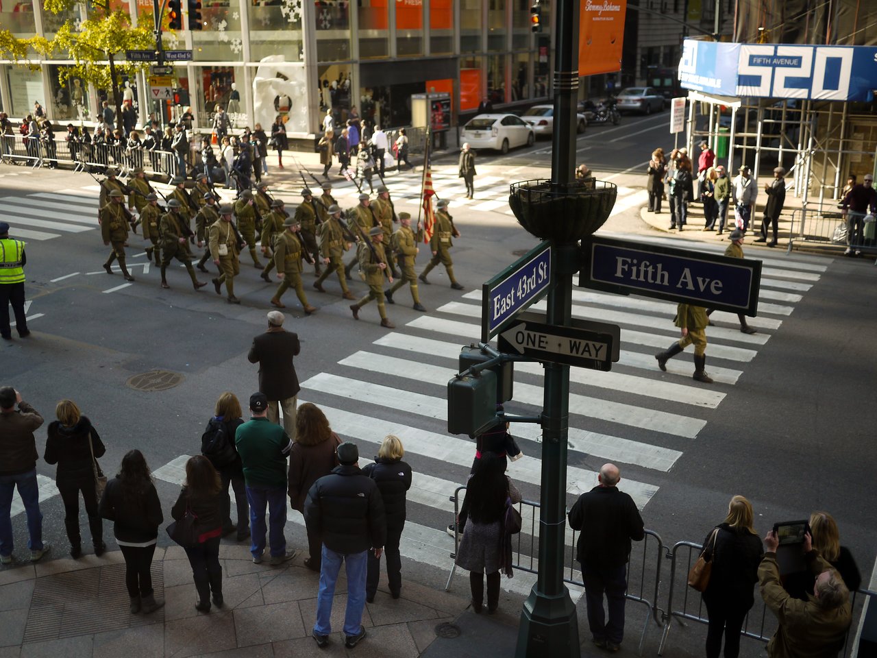Spectators at Fifth Avenue and East 43rd Street watch marchers in olive World War II style uniforms cross the street.