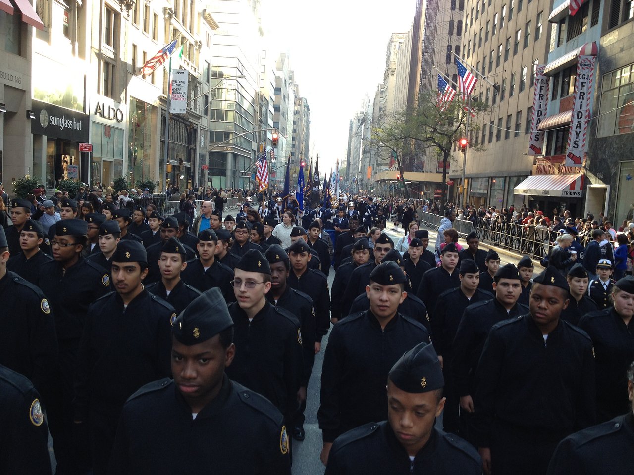 Young cadets in black uniforms and caps fill Fifth Avenue during the Veterans Day parade, with spectators and flags along the street.