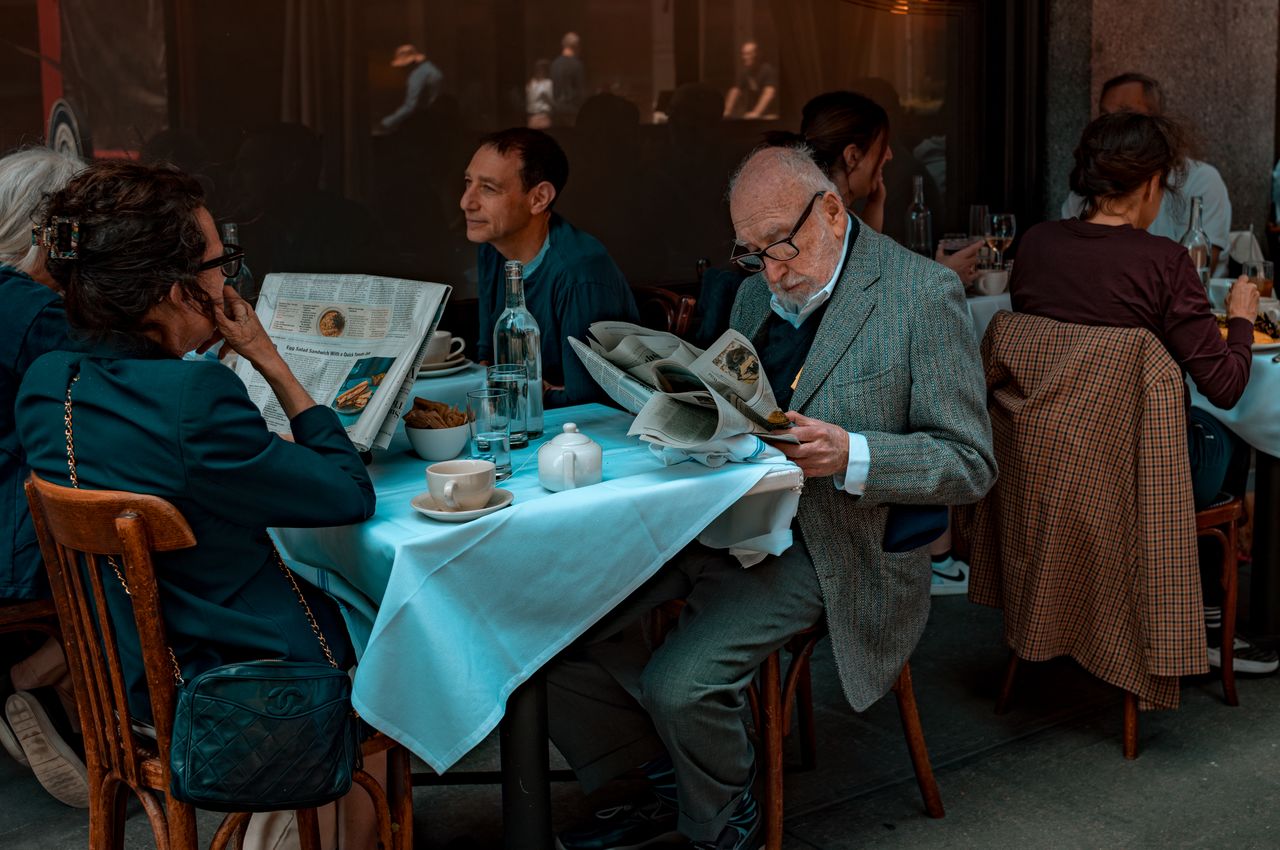 An older man in a gray suit reading a newspaper at an outdoor restaurant table.