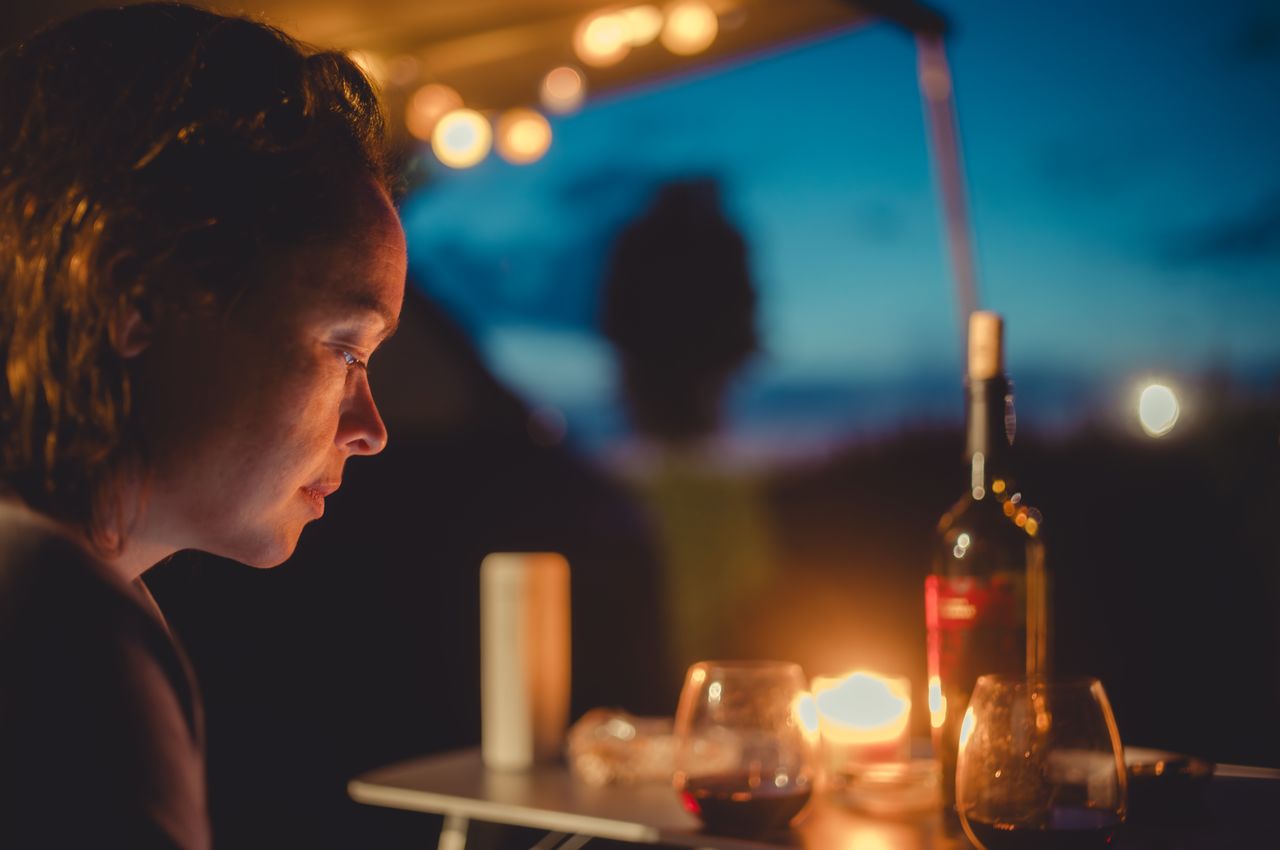 A woman sitting at a campsite table, lit by candlelight, with a bottle of wine and glasses.