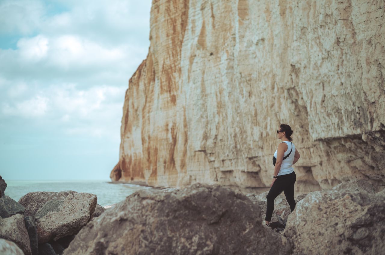 A woman stands on large rocks, looking at the sea with towering cliffs behind her.