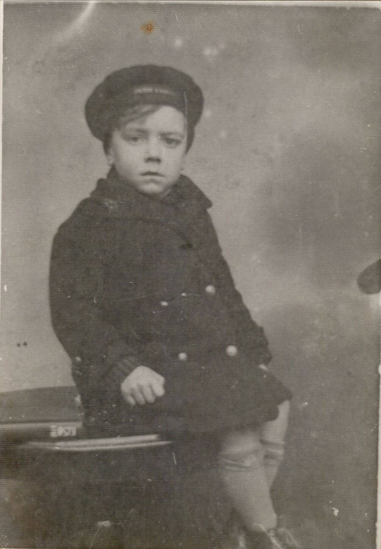 A young child wearing a dark coat and hat sits on a stool, looking seriously at the camera.