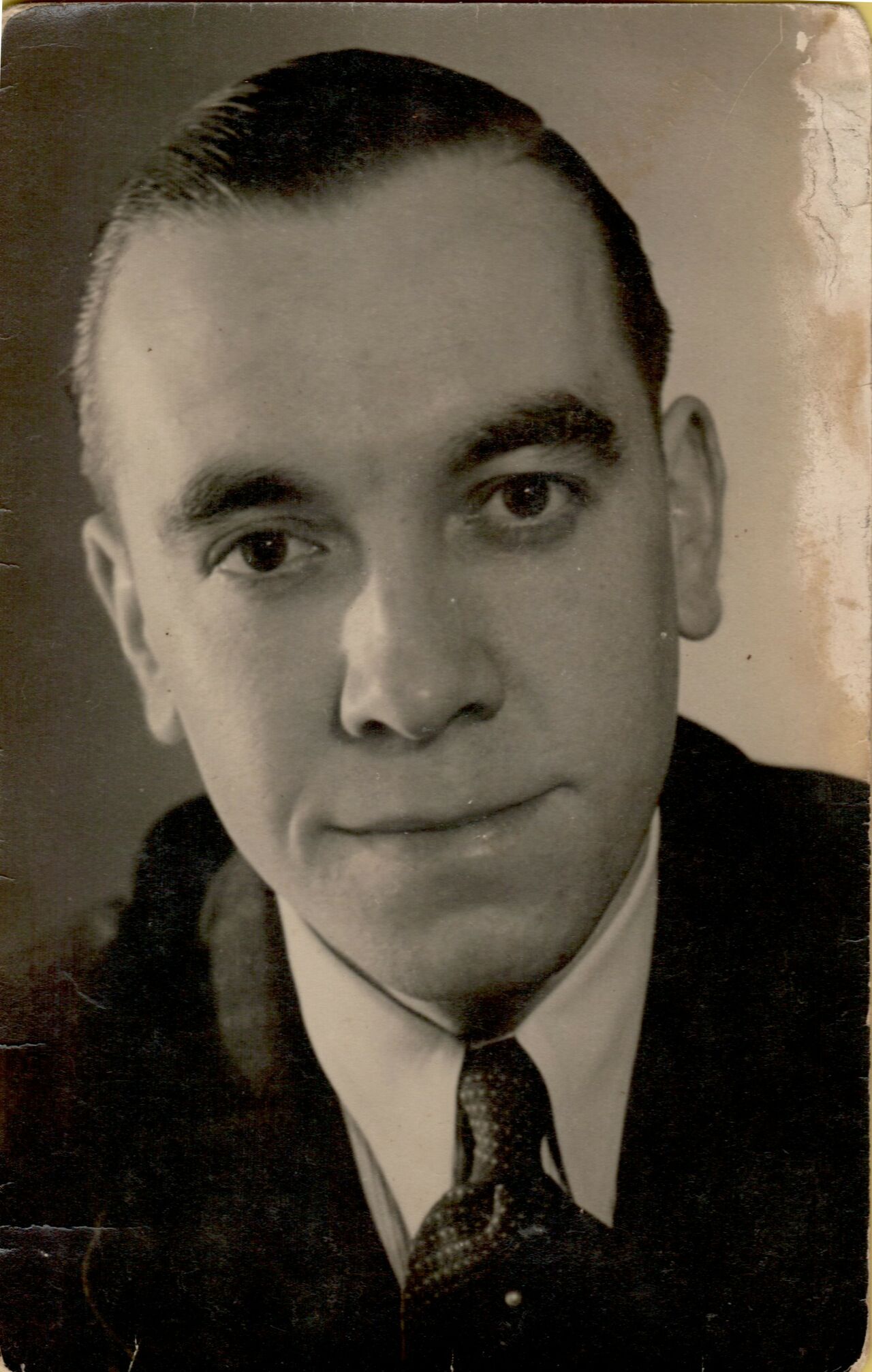 A black-and-white portrait of a man in a suit and tie, looking directly at the camera with a slight smile.