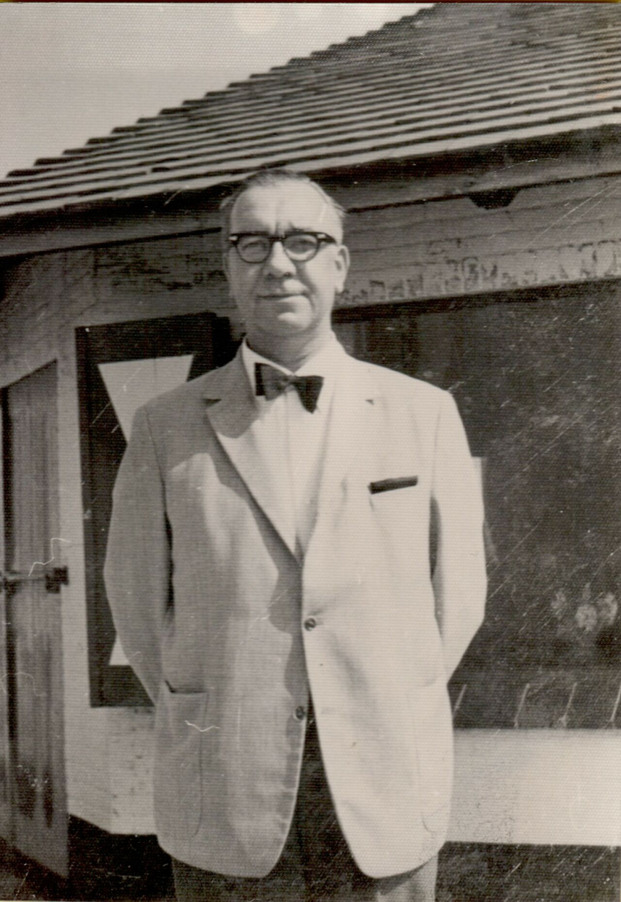 A man wearing glasses and a bow tie stands in front of a small building with a slanted roof.