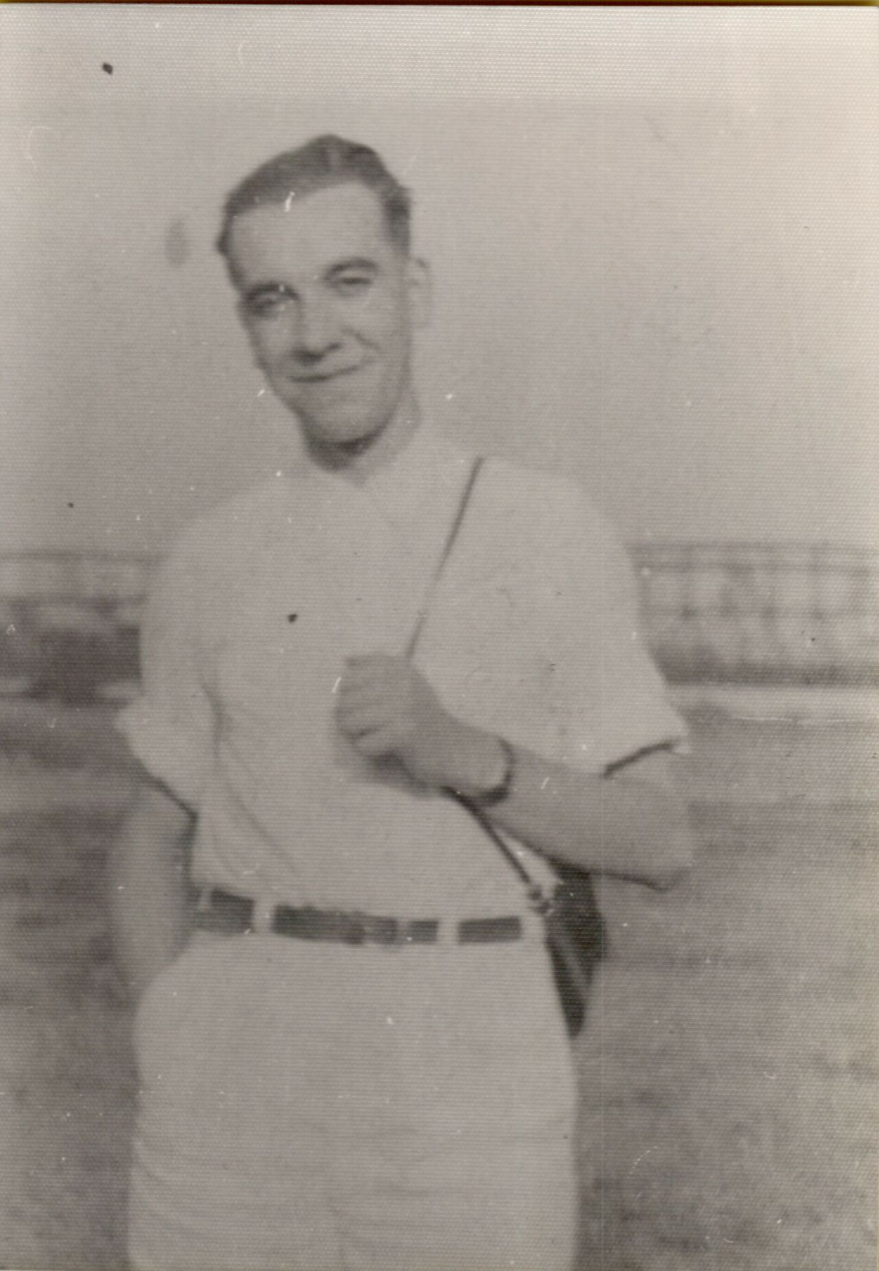 A man in a short-sleeved shirt and belt stands outdoors, smiling, with one hand in his pocket.