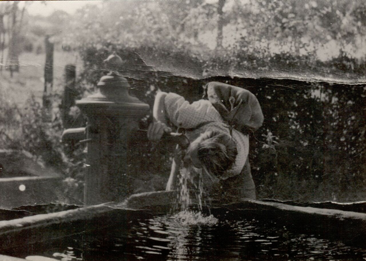 A young adult bends down to drink water from a fountain in an outdoor setting.