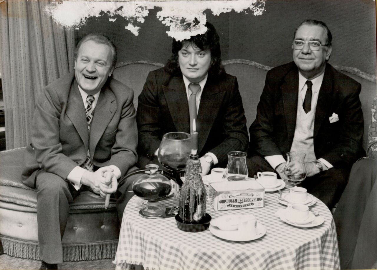 Three men in suits sit on a couch, with a box of Jules Destrooper cookies on the table in front.