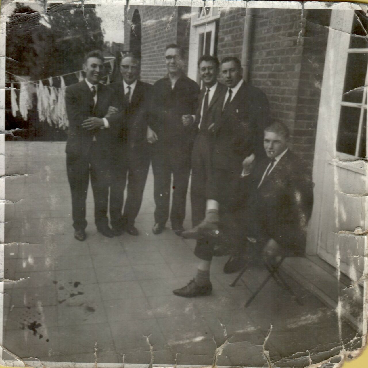 A group of seven men posing together outside a brick building, dressed in suits, with one playfully lifting his leg.