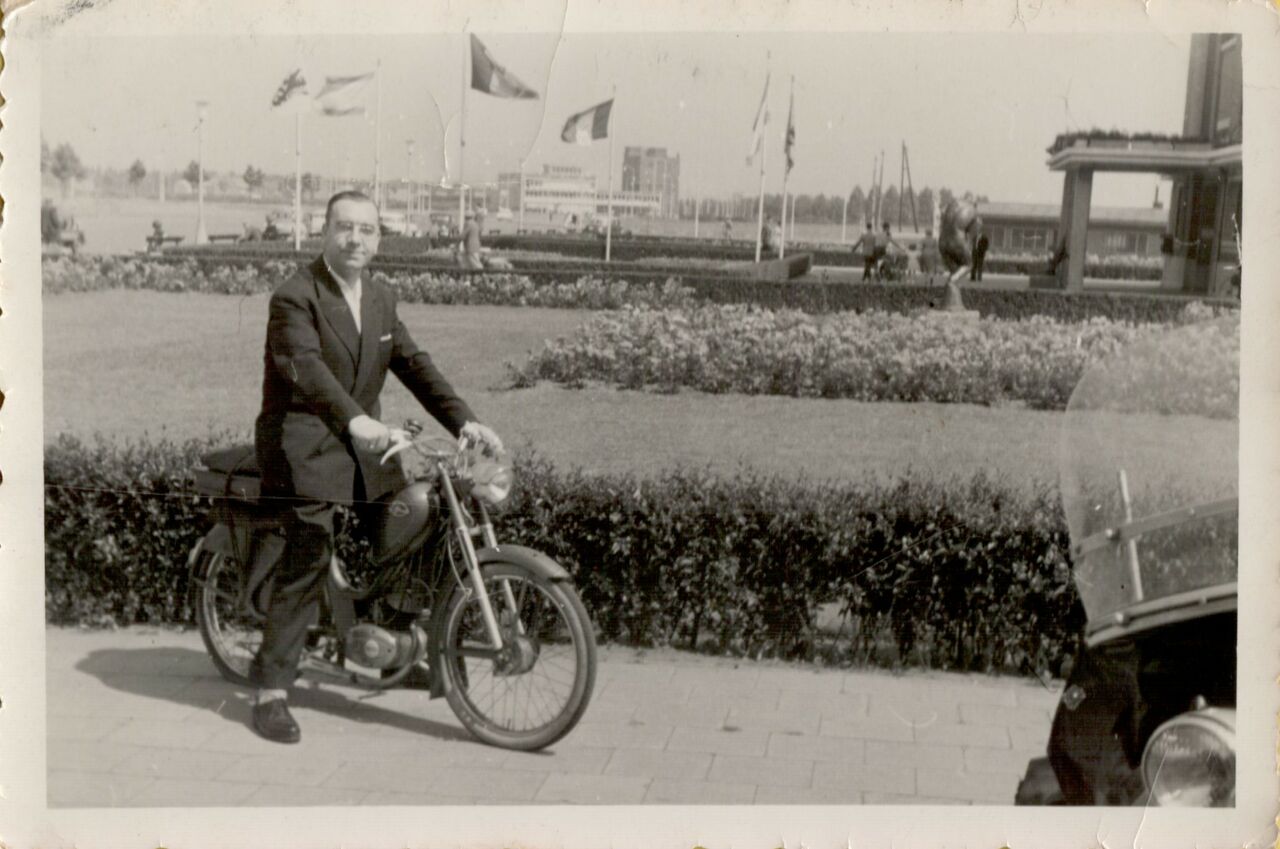 A man in a suit stands next to a Zundapp motorcycle on a paved path with flags in the background.