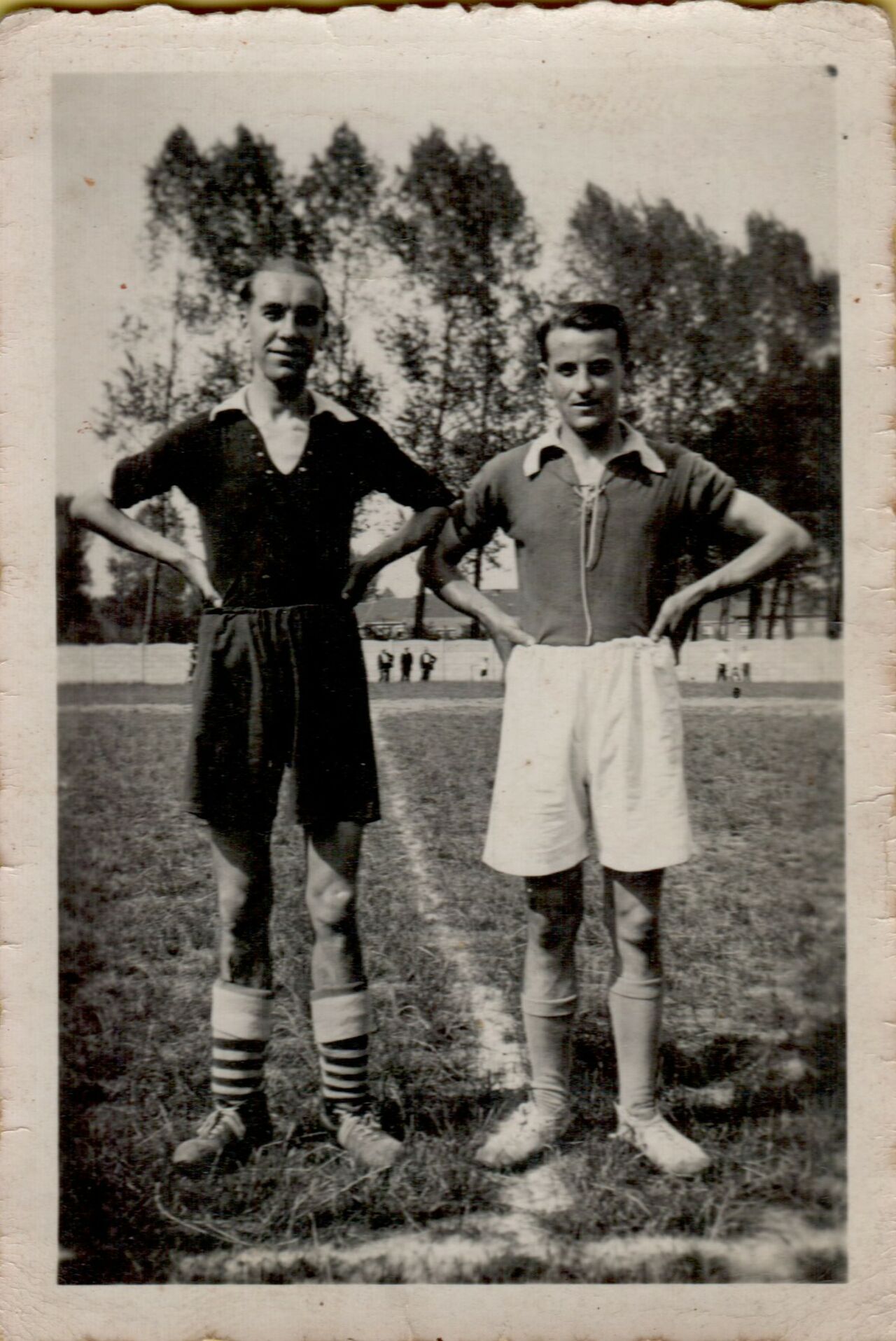 Two soccer players stand on a grass field with hands on hips, wearing vintage uniforms and striped socks.