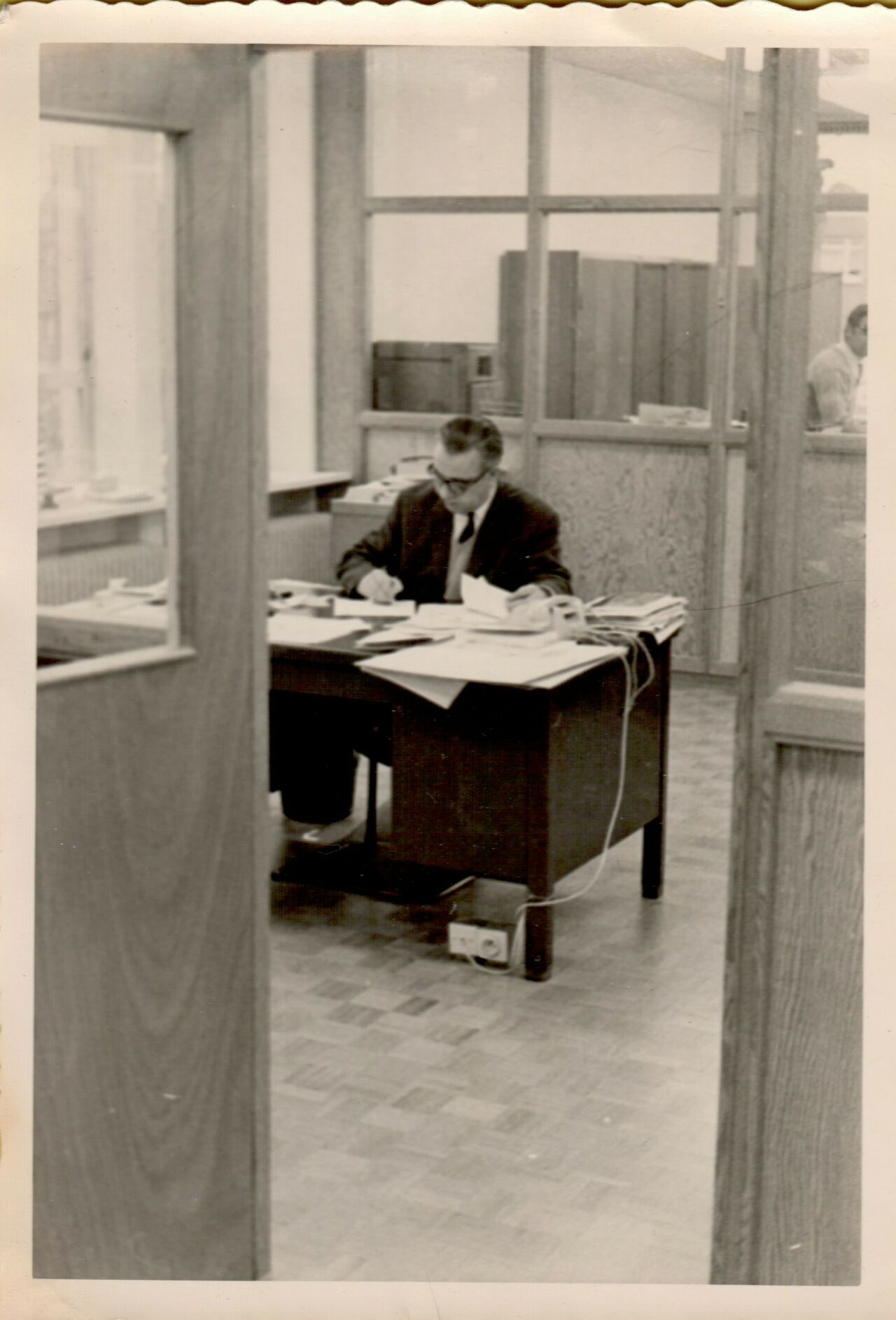 A man in a suit sits at a desk, writing on papers in an office with glass partitions.