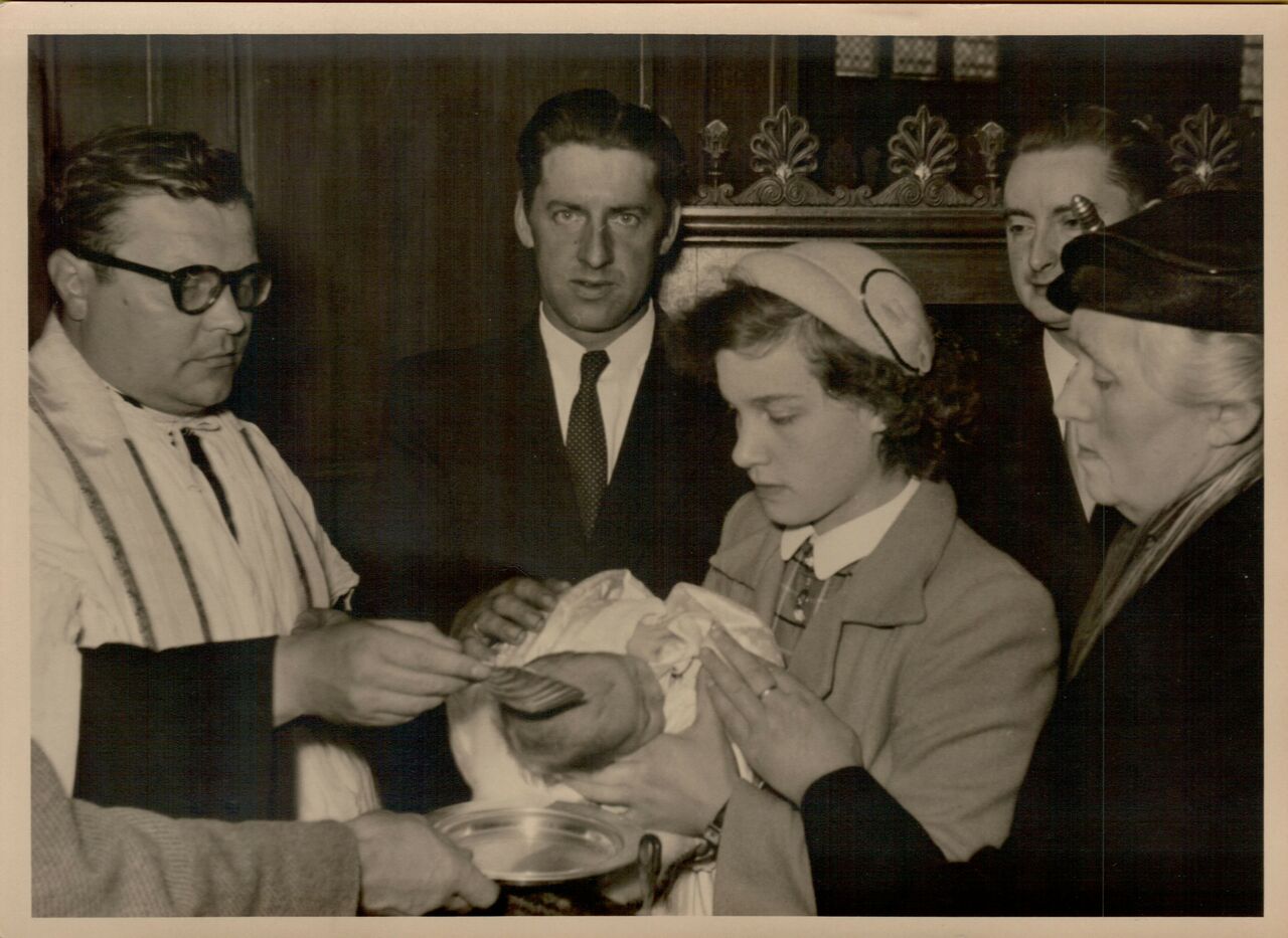 A priest performs a baptism as a woman holds the baby, surrounded by family members in a church setting.