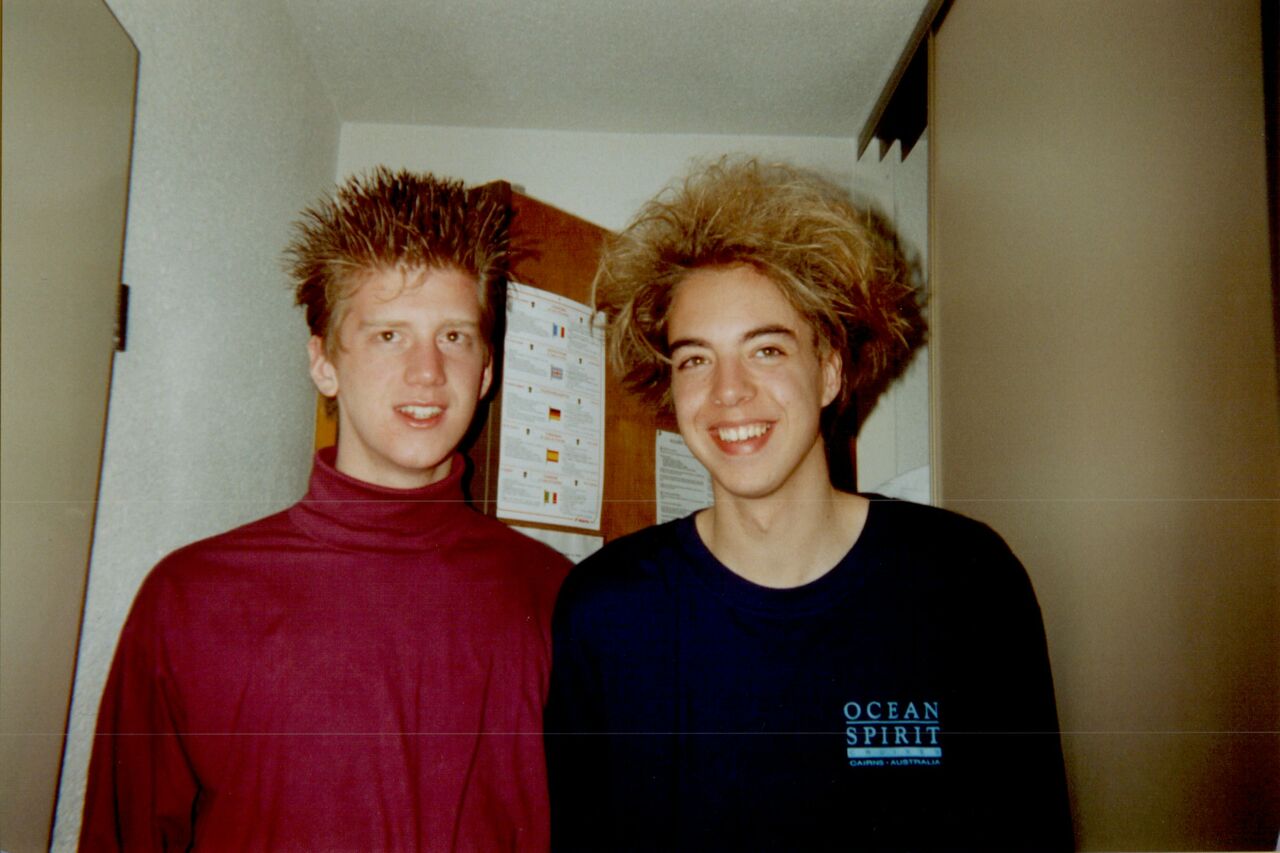 Two young men with spiky and frizzy hair smile at the camera in an indoor setting.