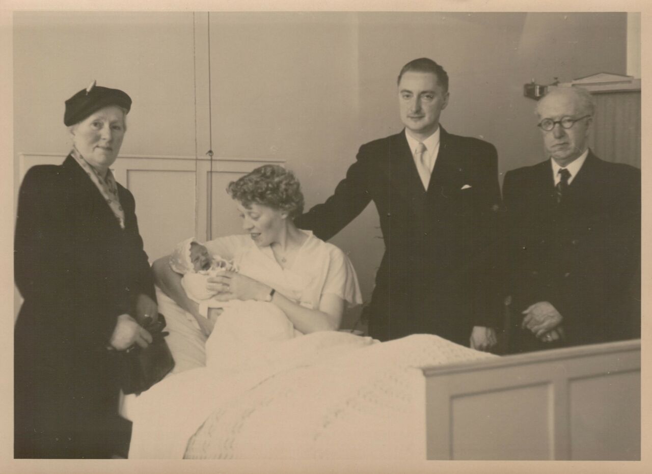 A mother in bed holds a newborn baby, surrounded by three formally dressed adults in a hospital room.