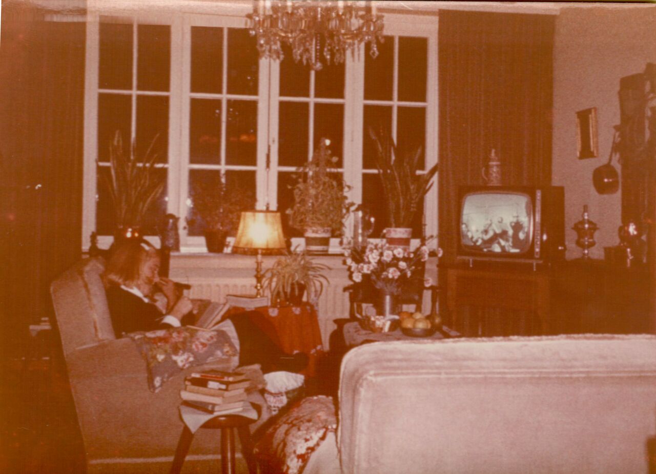 An elderly person sits in an armchair reading and smoking a pipe, while a television plays in the background.